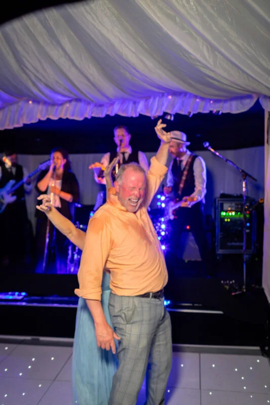 A joyful wedding guest dances enthusiastically on a lighted dance floor under a marquee ceiling while a live band performs on stage in the background.