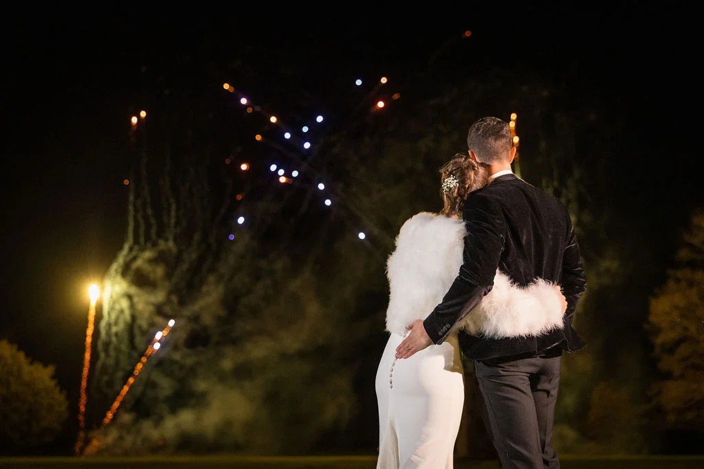 A newlywed couple embraces from behind while watching a vibrant fireworks display light up the night sky during their evening wedding reception.