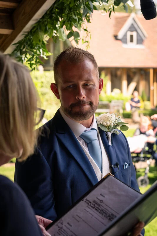 An emotional groom dressed in a navy suit and light blue tie listens intently during an outdoor wedding ceremony.