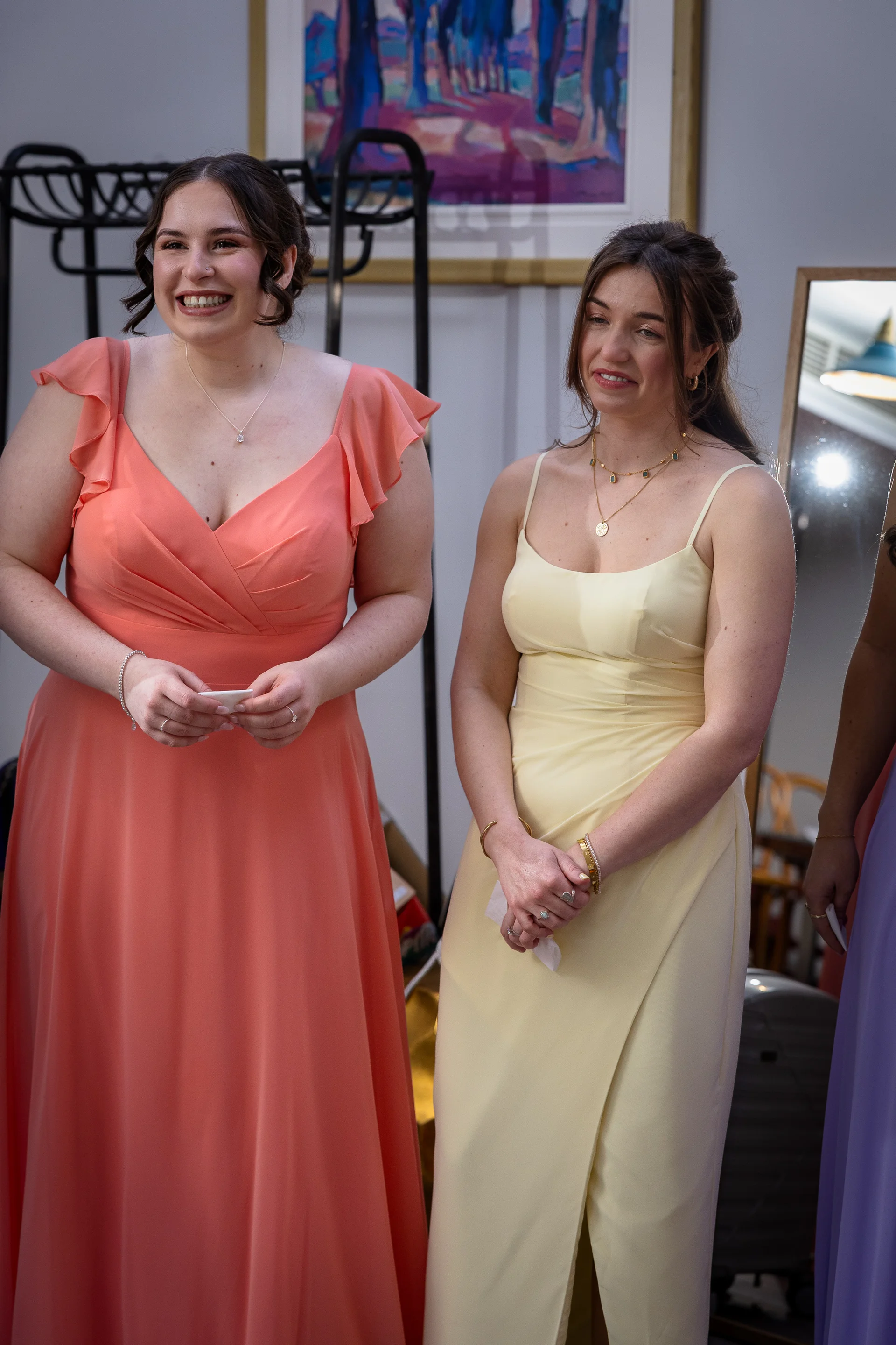 Two smiling bridesmaids stand side by side wearing pastel dresses in peach and soft yellow during a joyful wedding moment.