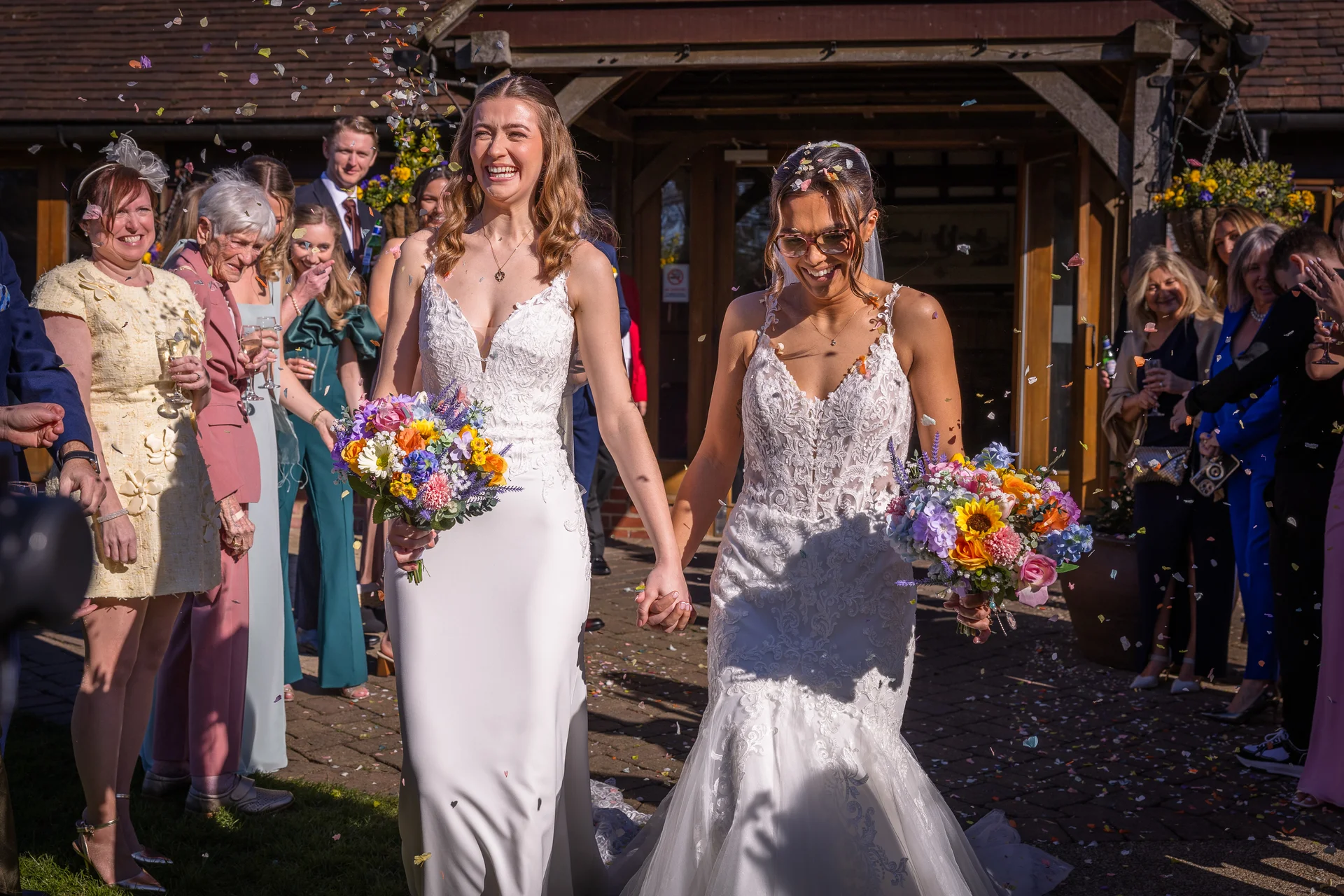 Two smiling brides holding hands and walking through a shower of colorful confetti outside a rustic stone barn venue on a sunny day.