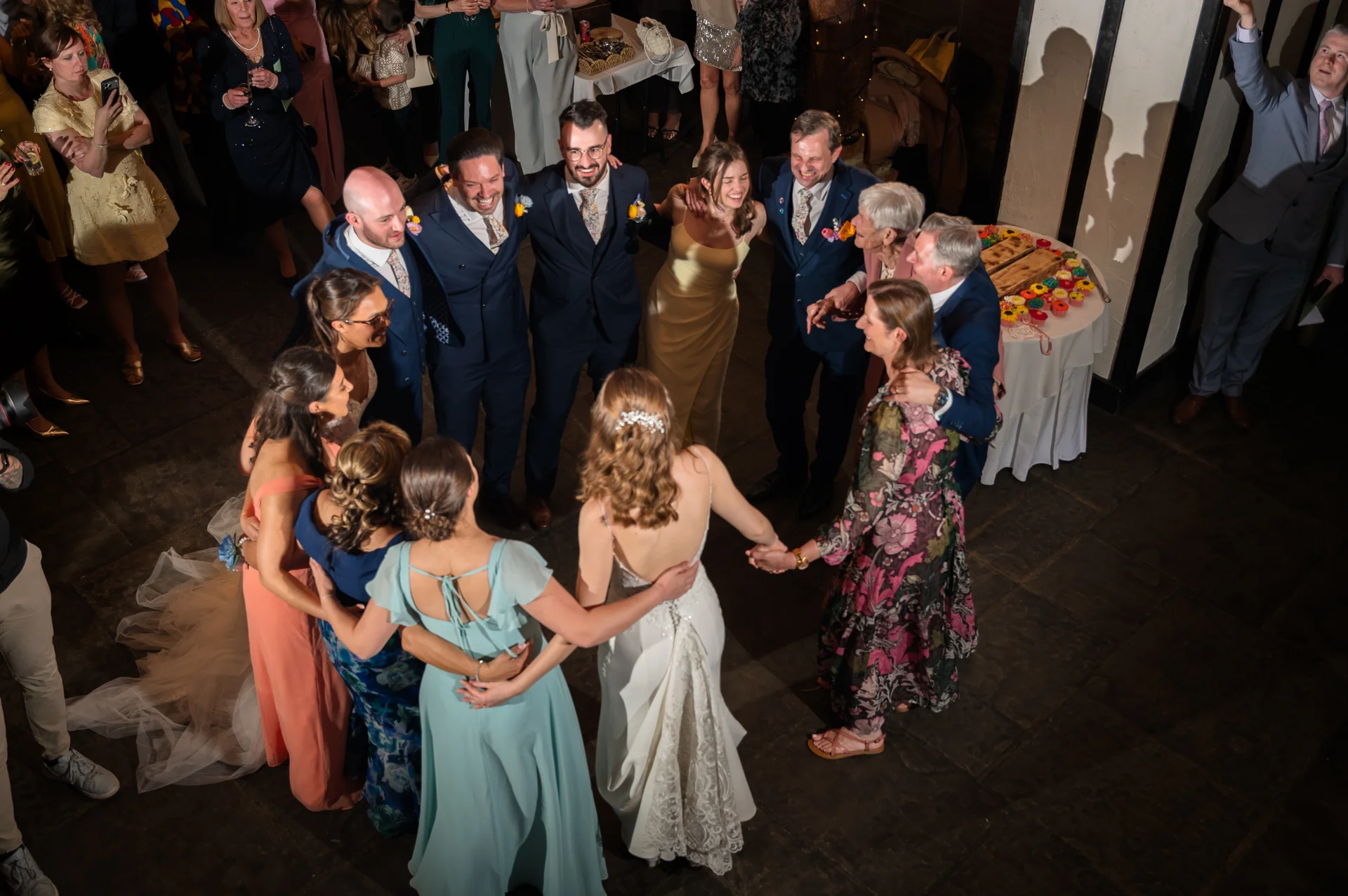 An aerial view captures a joyful wedding party and guests forming a large circle on the dance floor during the evening reception.