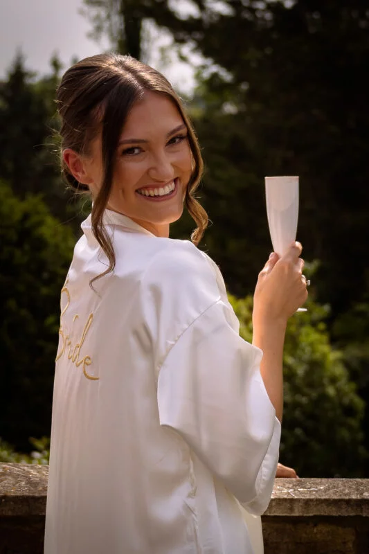 A smiling bride wearing a white satin robe with 'Bride' embroidered on the back looks over her shoulder while holding a champagne flute.