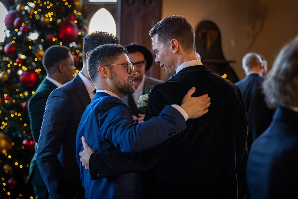 Two grooms in formal suits embrace warmly during a festive winter wedding celebration, surrounded by guests and a glowing Christmas tree.