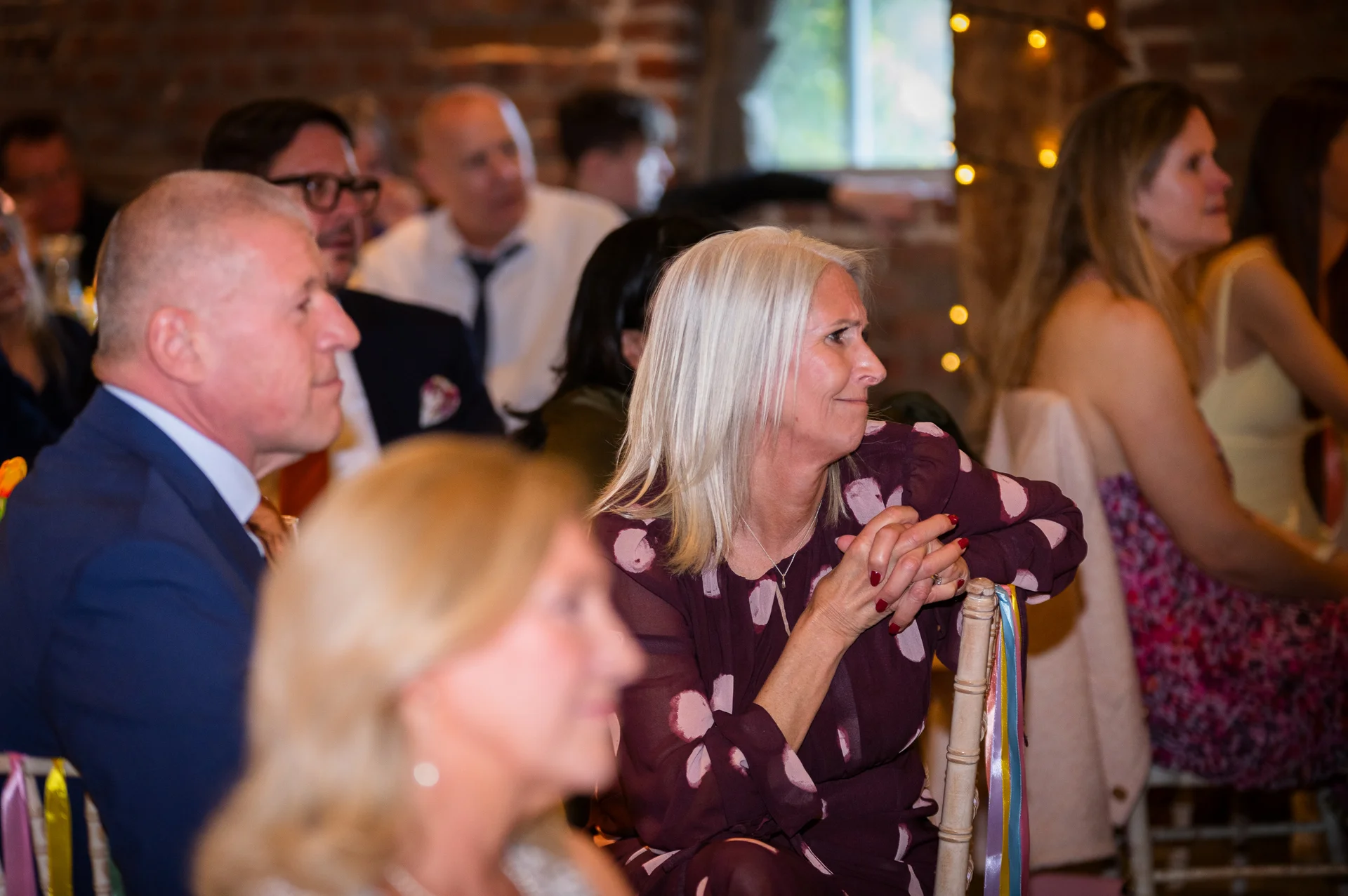 Wedding guests seated indoors at a reception, watching attentively with warm, natural expressions in an exposed brick venue.
