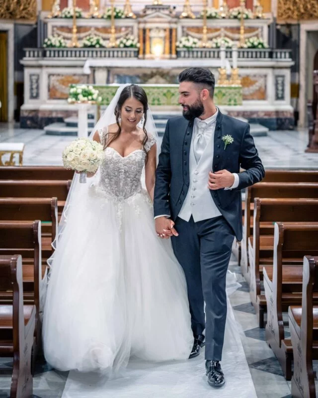 A newlywed couple smiling and walking hand in hand down the aisle of a grand church following their wedding ceremony.