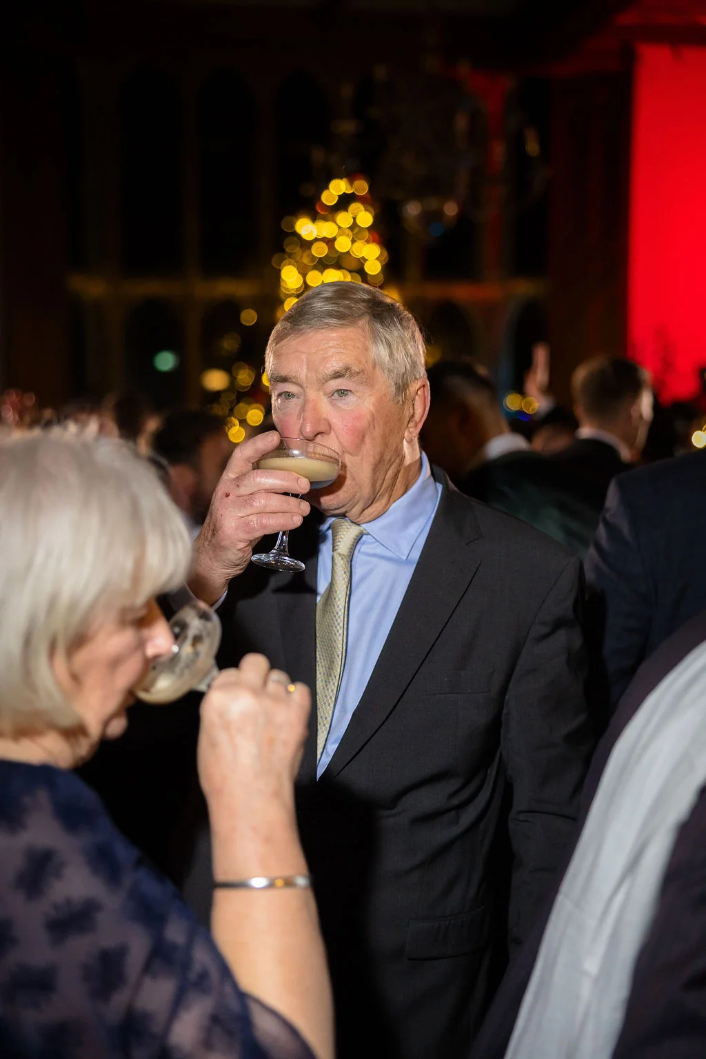 An older gentleman enjoys a cocktail from a coupe glass while mingling with guests at a lively evening wedding reception.