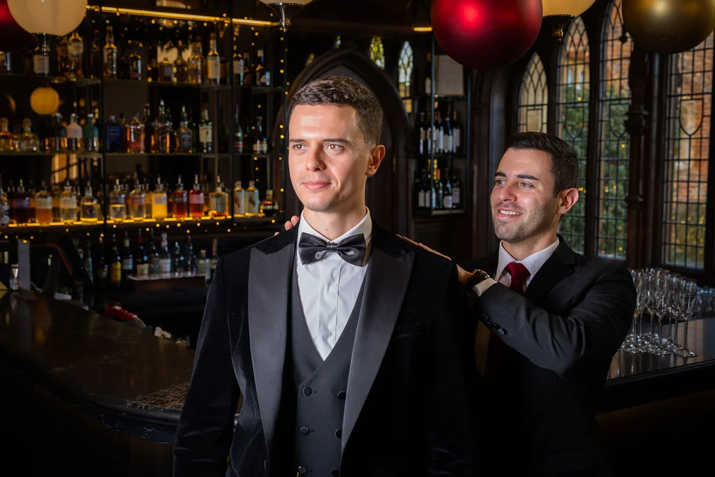 One groom in a black velvet tuxedo and waistcoat stands proudly as his partner gently places a hand on his shoulder in a sophisticated bar setting.