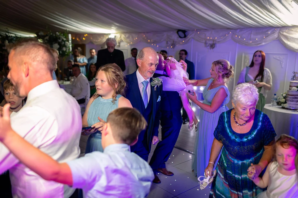 Guests of all ages enjoying the evening reception on a lit dance floor inside a draped marquee with ambient purple lighting.
