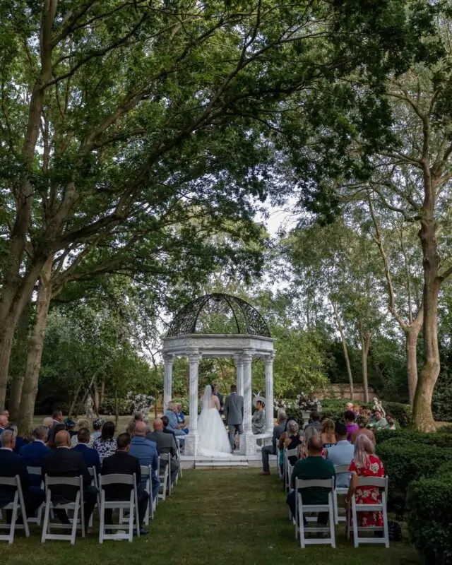 An outdoor wedding ceremony takes place inside a stone gazebo nestled beneath a lush canopy of mature trees with seated guests watching.
