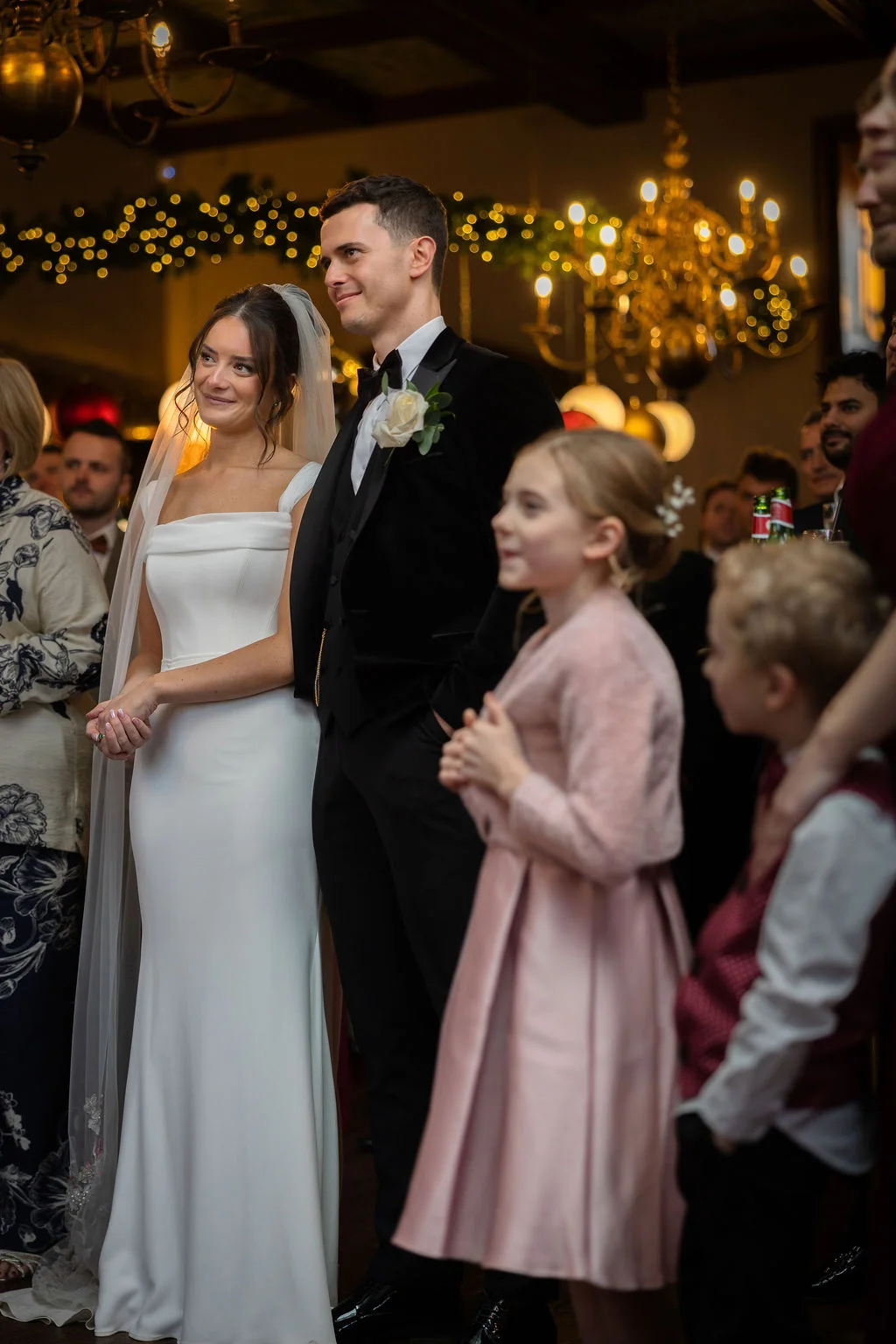 A newlywed bride in a sleek white gown and veil stands beside her groom in a velvet tuxedo during a candlelit evening reception.