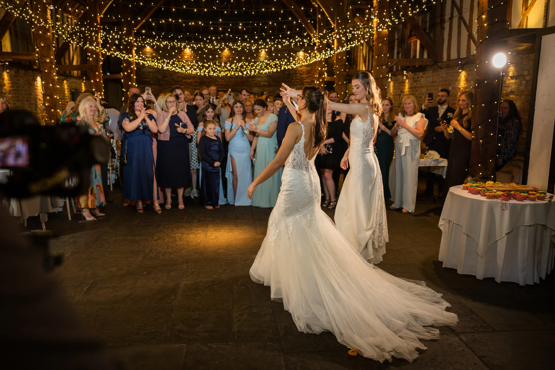 Two brides share their first dance in a rustic barn venue illuminated by a warm fairy light canopy while guests watch and applaud.