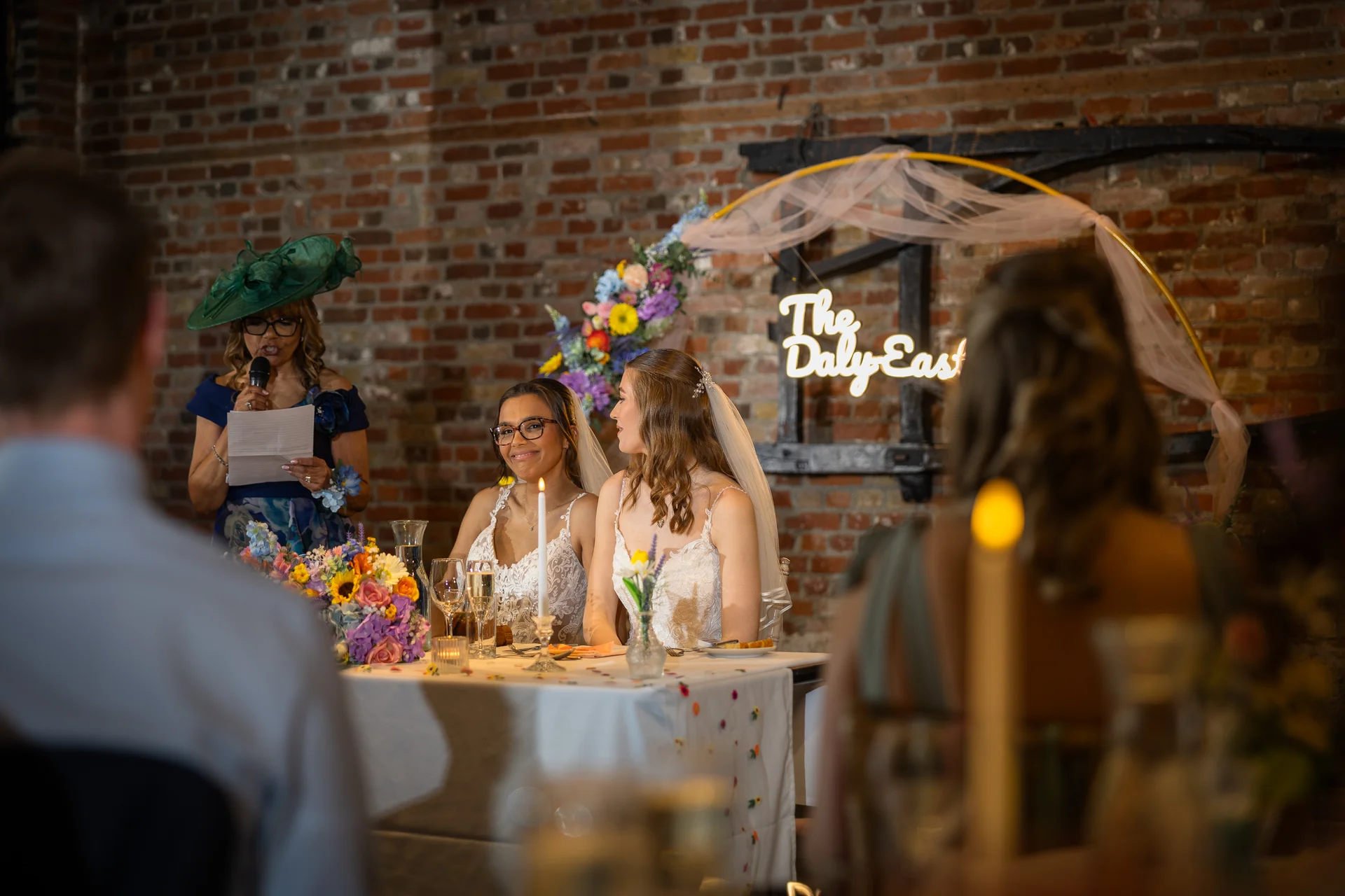 Two smiling brides sit together at the top table during wedding speeches in a rustic exposed brick venue while a guest gives a toast.