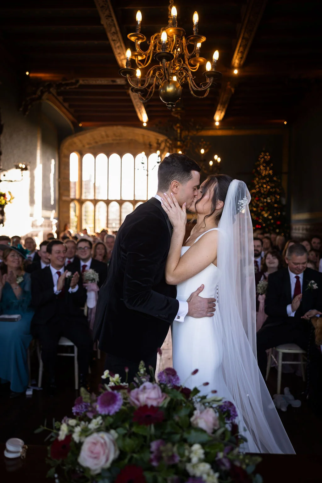 Newlyweds share their first kiss at the altar inside a grand candlelit hall with an ornate chandelier and applauding guests watching.