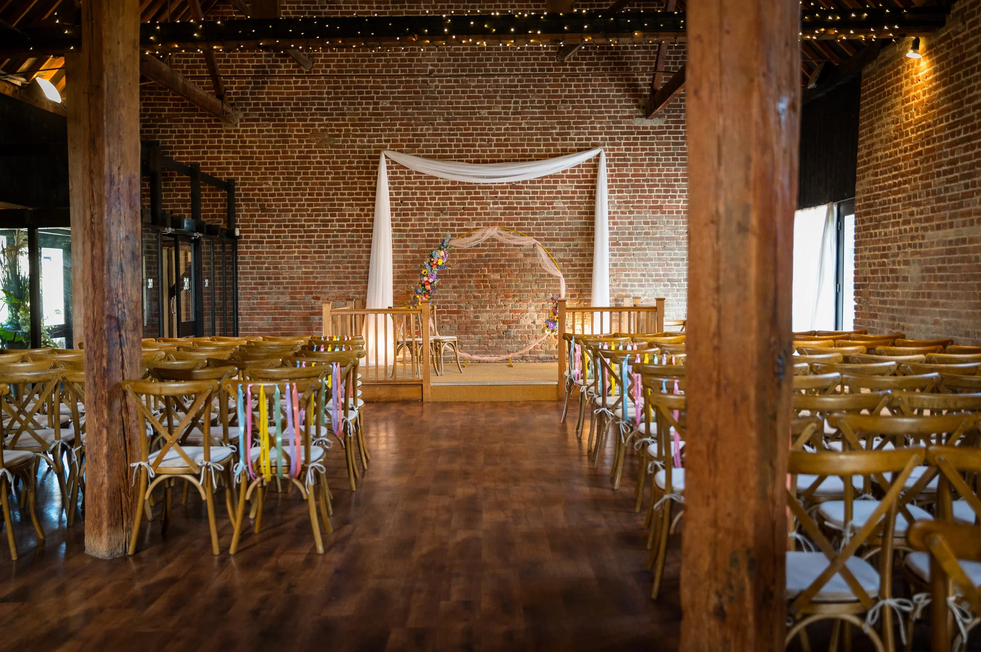 A rustic barn wedding ceremony setup featuring exposed brick walls, wooden cross-back chairs adorned with colourful ribbons, and a circular floral arch at the altar.
