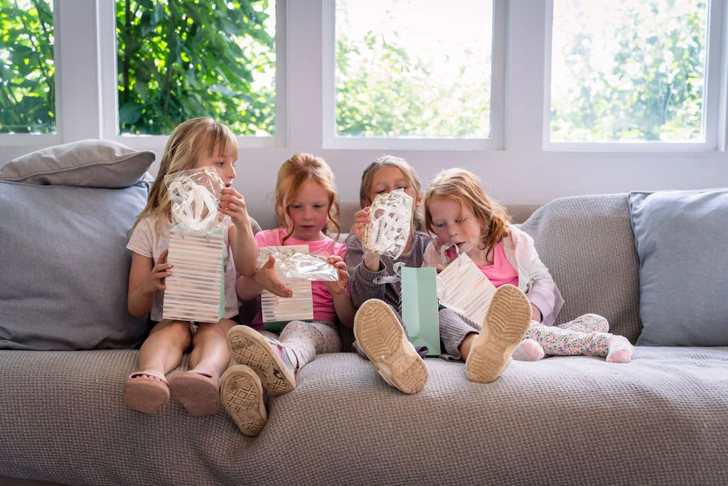 Four young flower girls sit together on a grey sofa, eagerly opening small gift bags in natural window light.