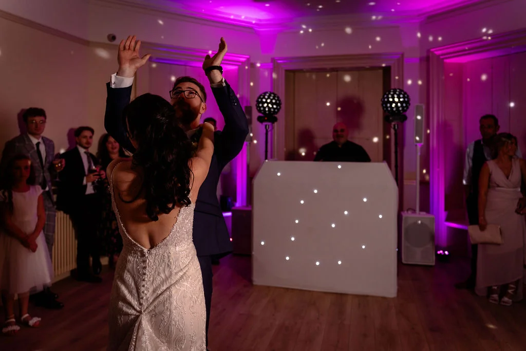 The newlywed couple shares their first dance with arms raised under vibrant purple disco lighting, surrounded by watching guests and a DJ booth.