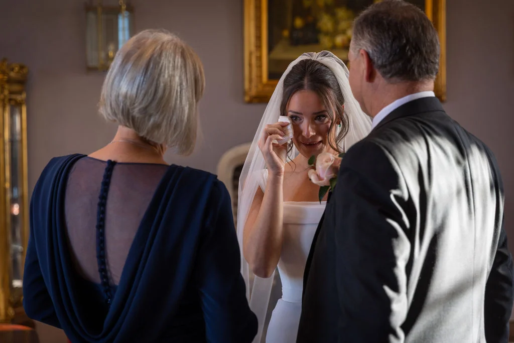 An emotional bride wipes away happy tears with a tissue while standing between her mother and father inside an elegant venue.