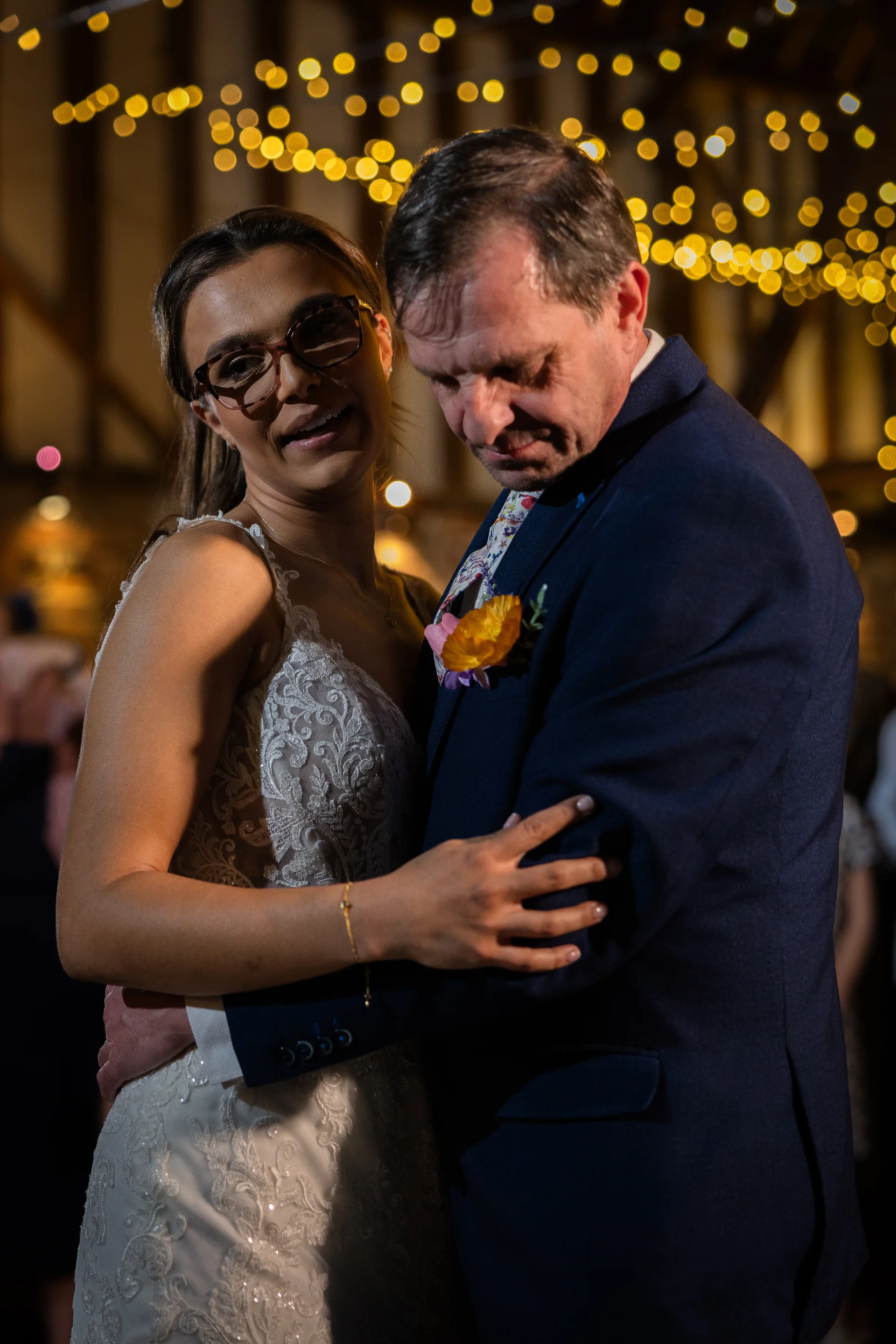 A newlywed couple embraces lovingly during their first dance under a warm canopy of fairy lights inside a rustic barn venue.