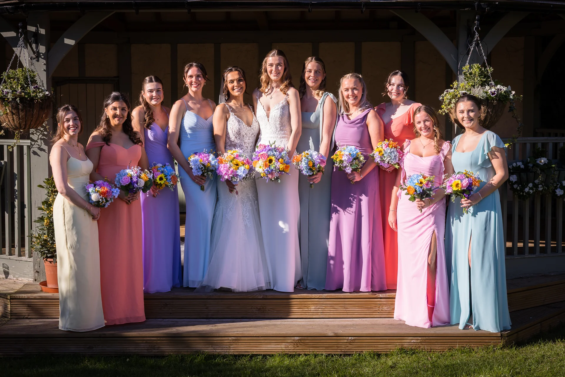 A bride and her large bridal party stand outdoors in the sunlight on wooden steps, holding colourful bouquets and wearing mismatched pastel dresses.