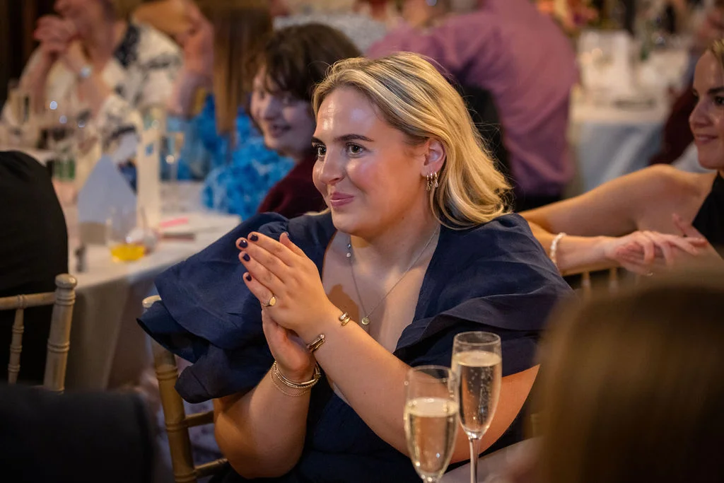 A smiling wedding guest in a dark blue dress claps happily at her table during the wedding reception speeches.