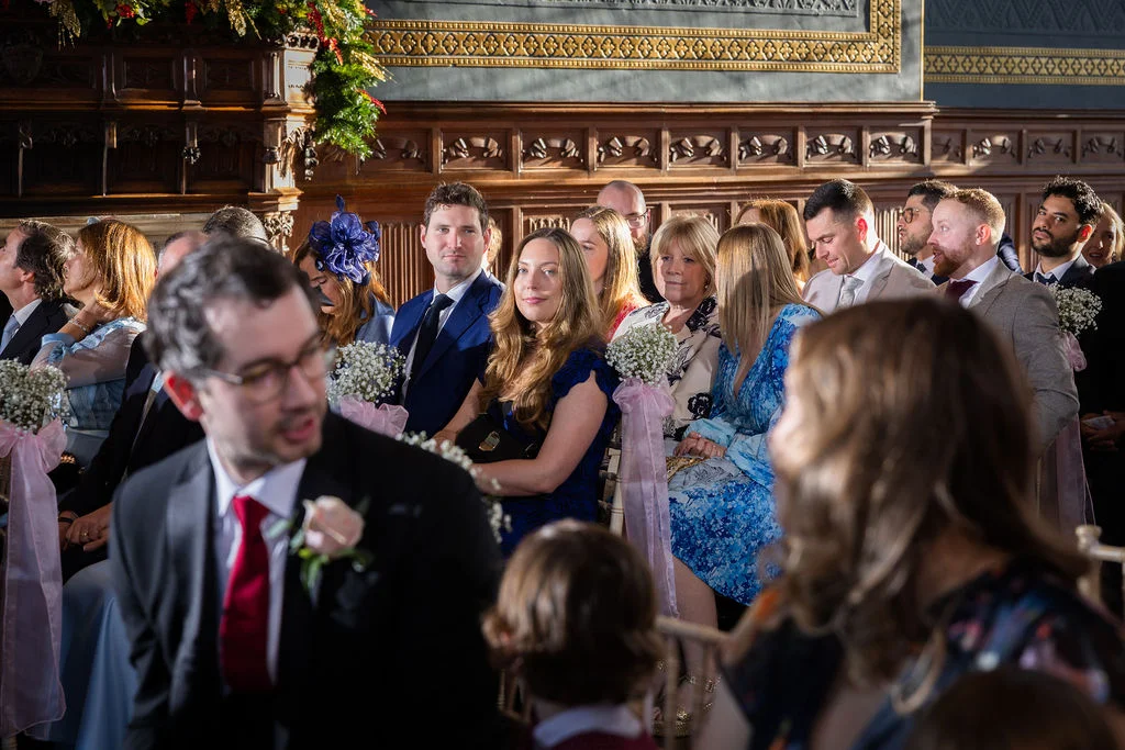 Wedding guests sit in a wood-panelled hall watching the ceremony, with a woman in the middle looking towards the camera while wearing a blue dress.