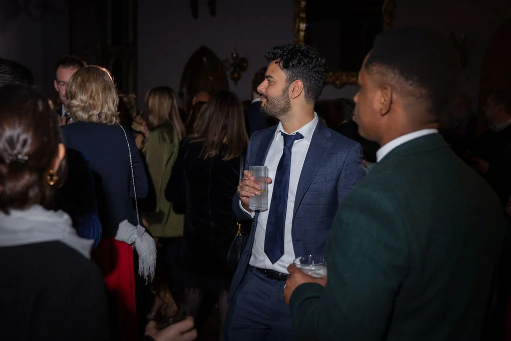 Dressed guests mingle and converse during an evening drinks reception, holding glasses in a softly lit venue space.