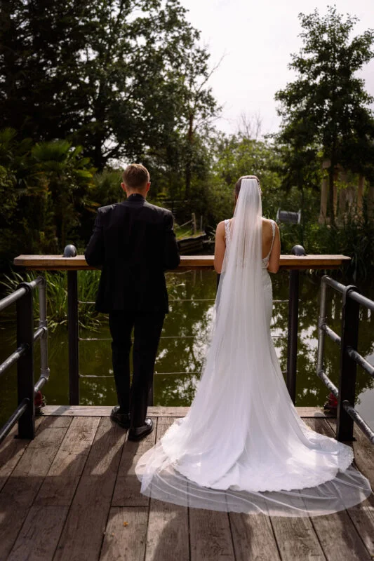 A bride and groom standing on a bridge overlooking a pond.