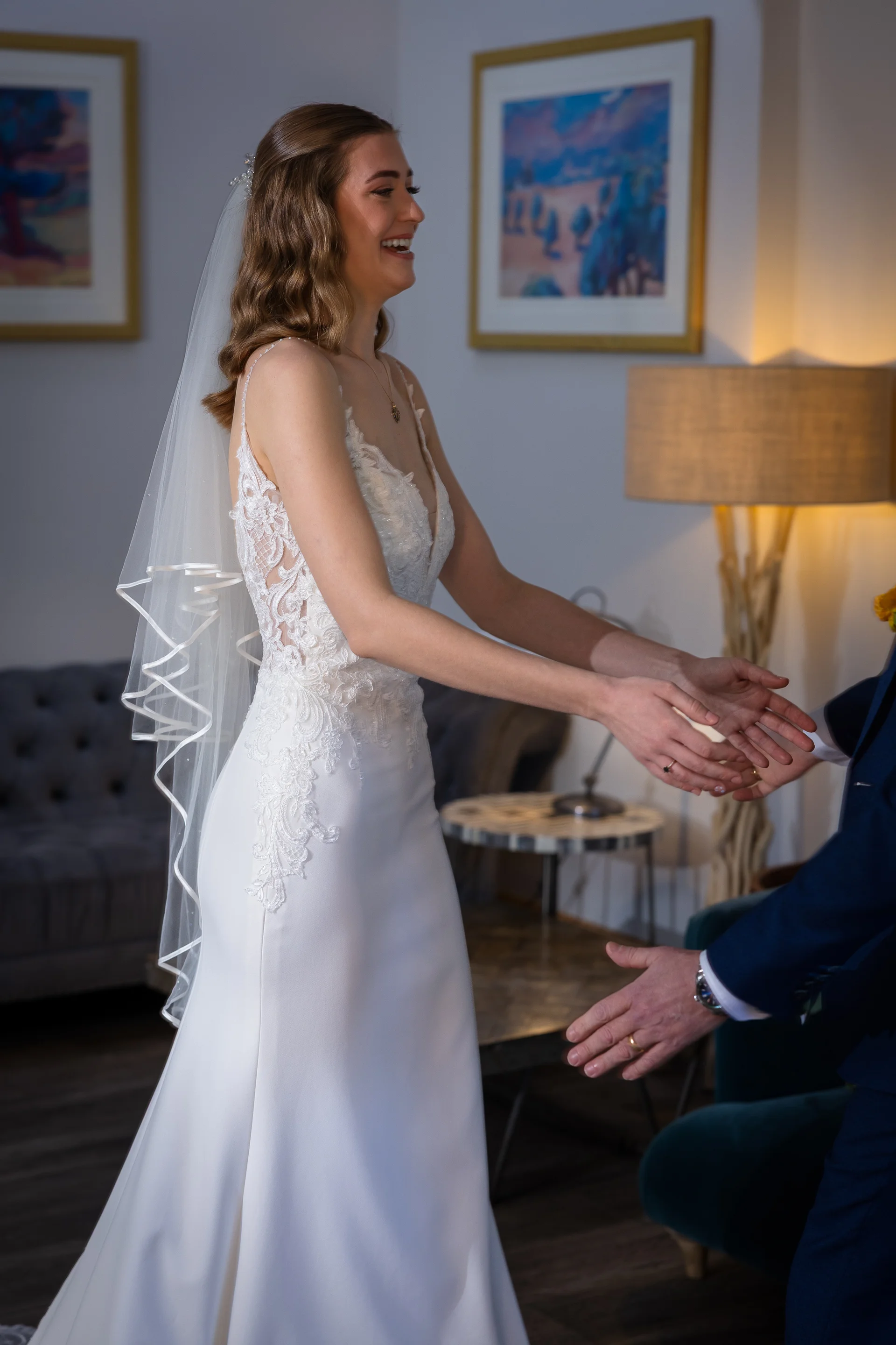 A joyful bride wearing a lace-detailed sheath gown and an elbow-length veil reaches out with a glowing smile during a first look moment with her groom.