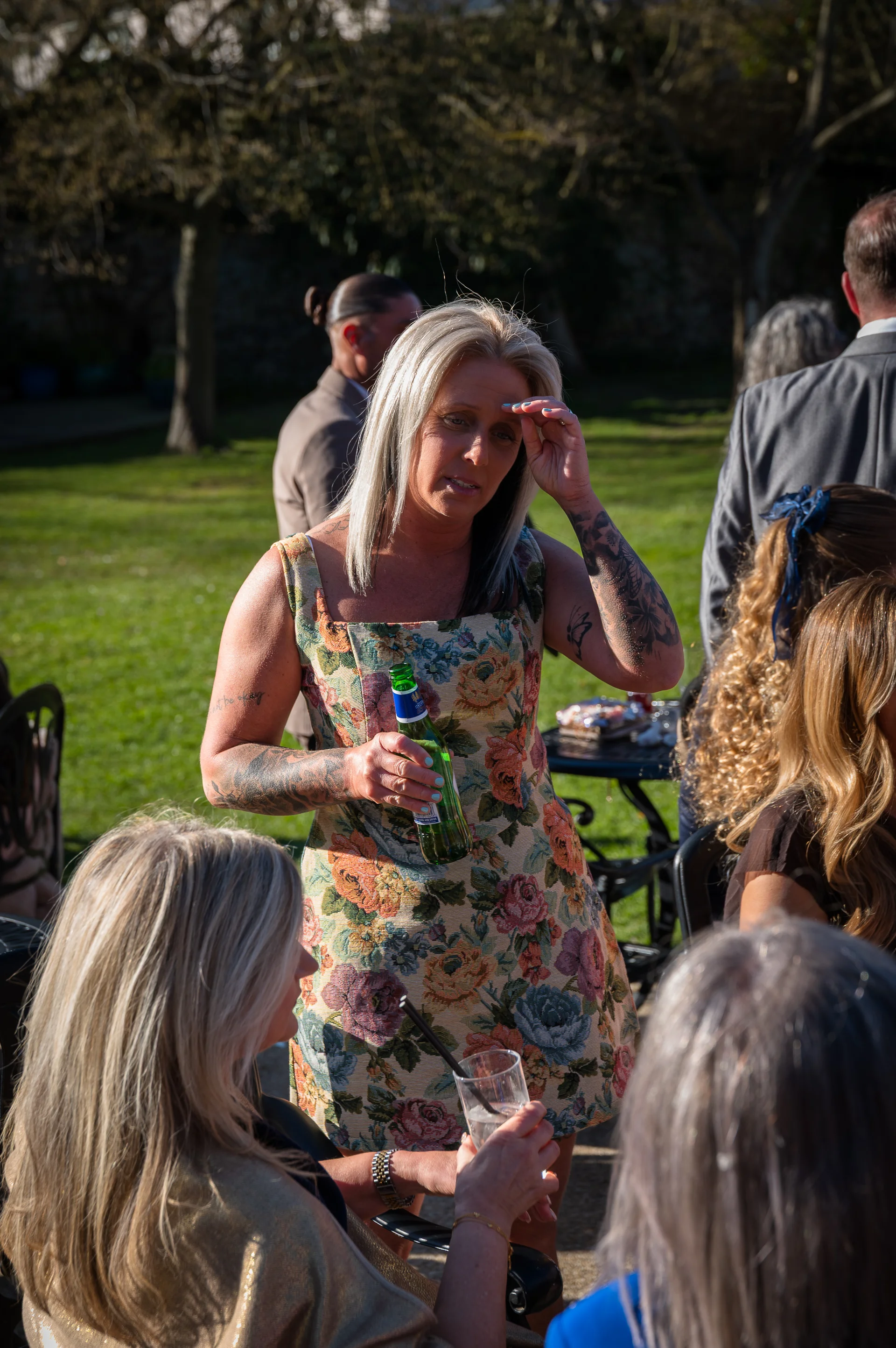 A blonde wedding guest wearing a floral print dress and holding a glass bottle stands outdoors in the sunshine among mingling guests.