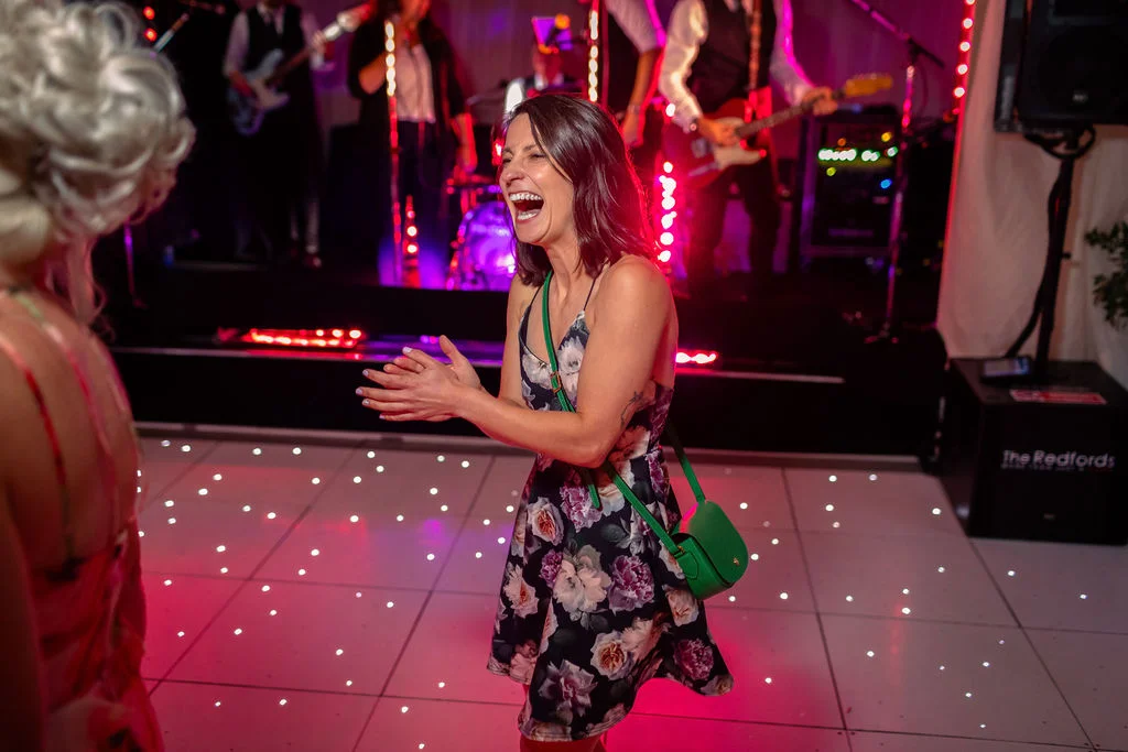 A wedding guest laughs joyfully while dancing on an illuminated dance floor in front of a live band performing on stage.
