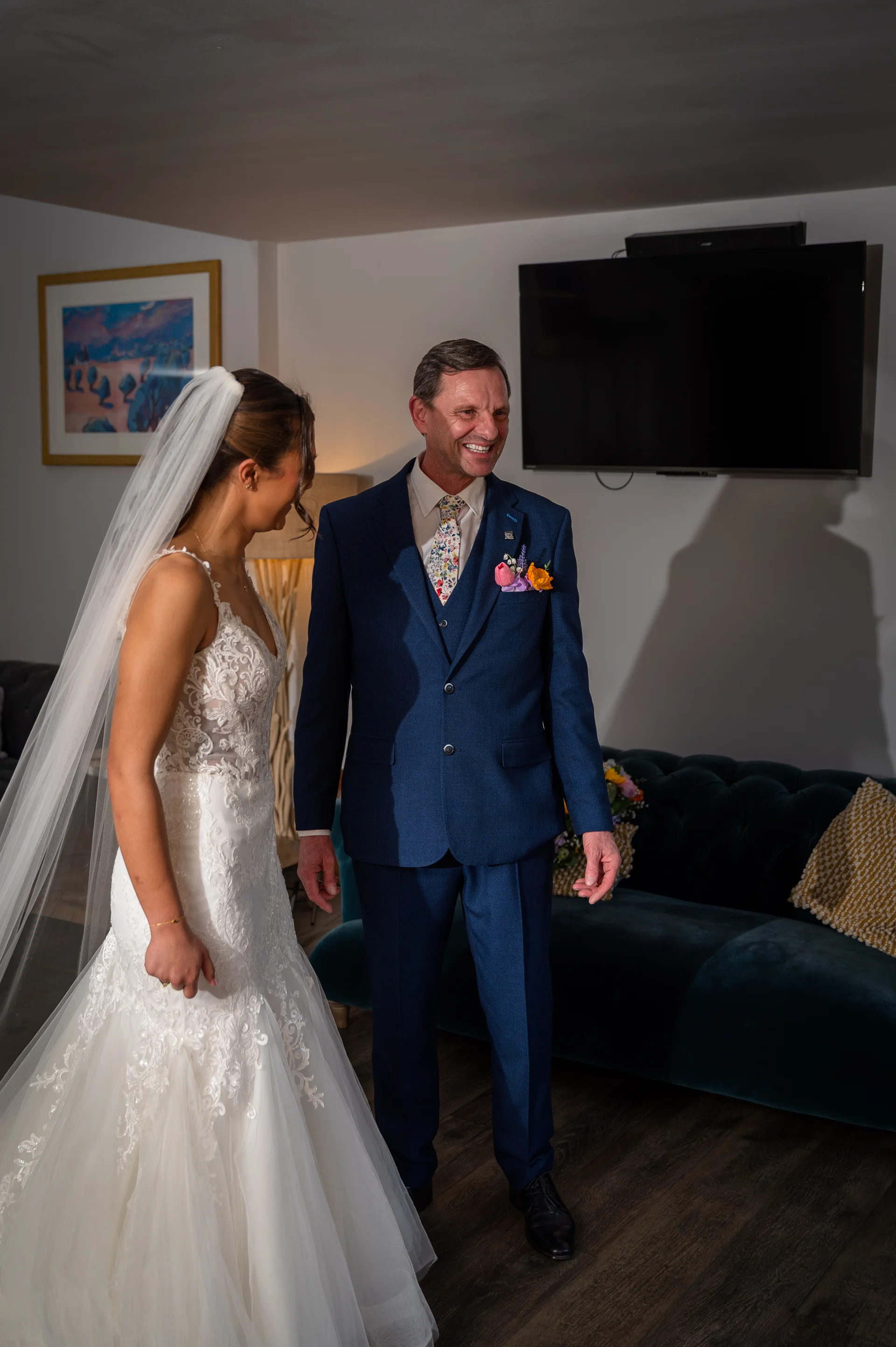 A beaming father of the bride in a smart blue suit shares a touching first look moment with his daughter in her lace wedding dress.