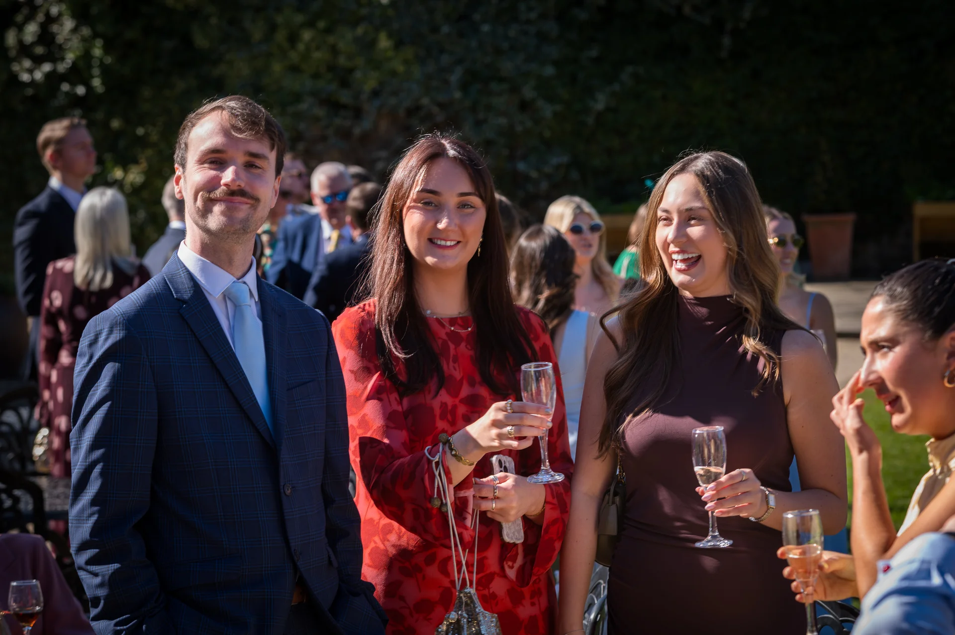 A group of smartly dressed wedding guests smile and laugh together outdoors in the sunshine while holding glasses of champagne during the drinks reception.