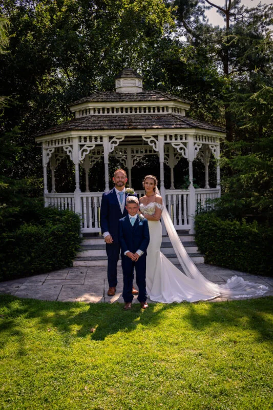 A newlywed couple and a young page boy pose together on a stone patio in front of a white wooden gazebo surrounded by lush green trees.