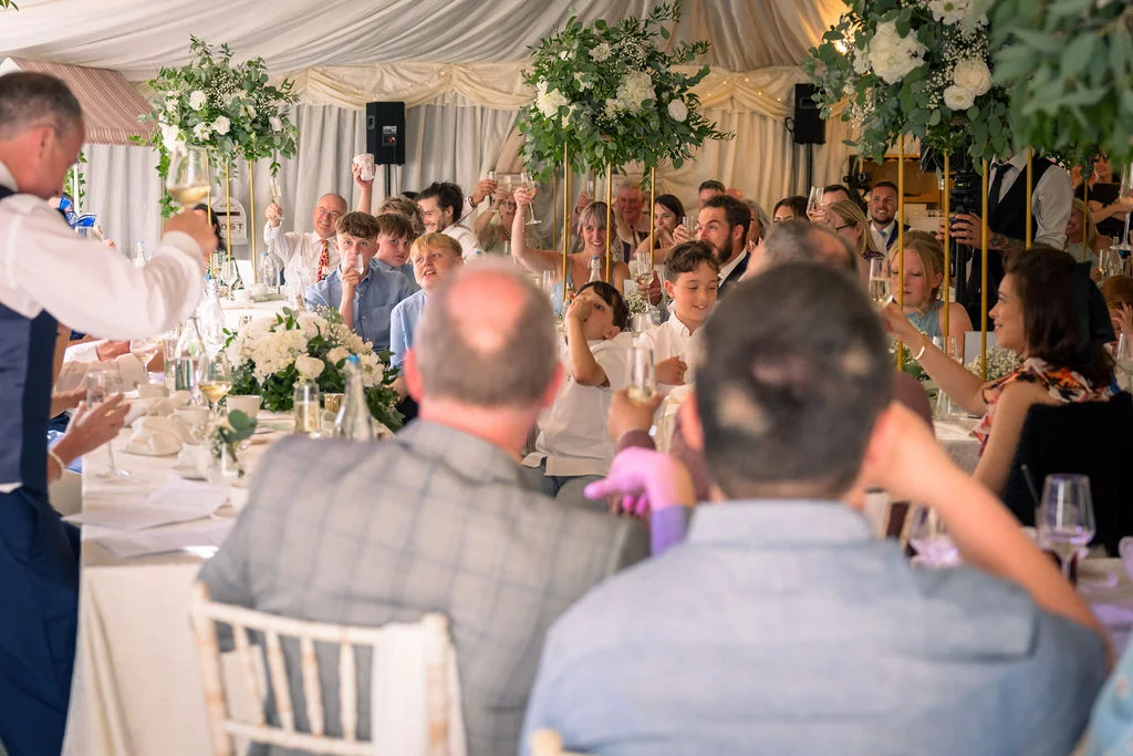Guests laugh and raise glasses during a wedding speech inside a brightly draped marquee decorated with tall floral centrepieces.