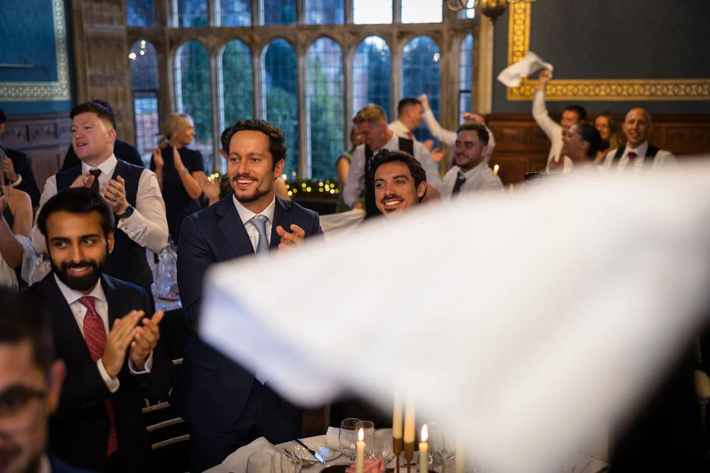 Wedding guests laugh and cheer enthusiastically while seated at tables during the wedding breakfast in a grand historic hall.