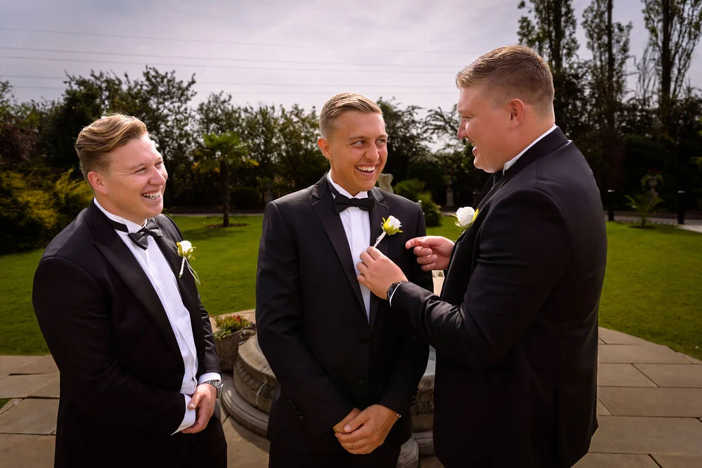 Three smiling groomsmen in black tuxedos share a joyful moment outdoors in a manicured garden while pinning a white rose buttonhole.