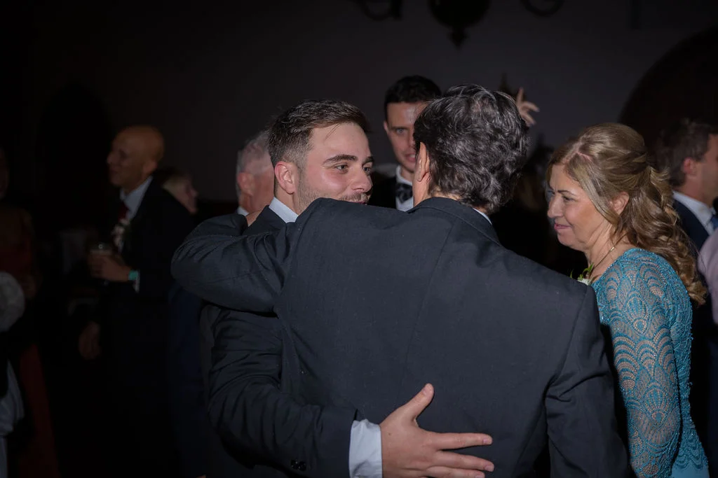 A guest and groom share a warm embrace during the wedding reception while other guests mingle in the background.