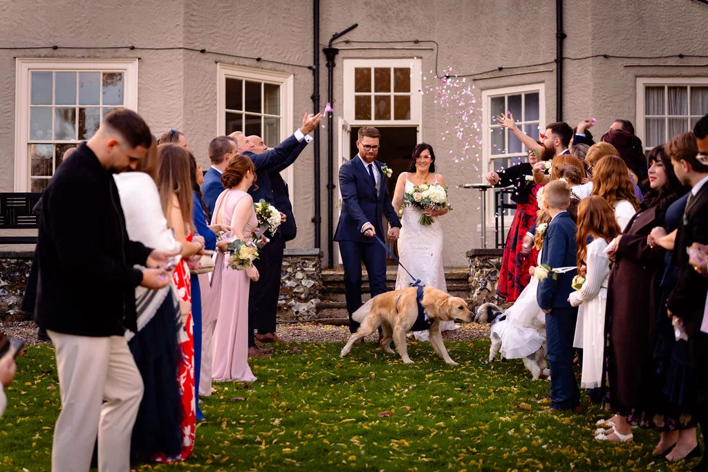 Newlyweds walk out of a country house hand in hand as guests shower them with confetti, accompanied by their golden retriever on a leash.