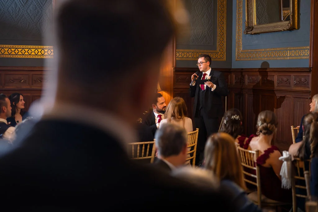 A groom delivers his speech while holding a microphone in an elegant wood-panelled room filled with seated guests.