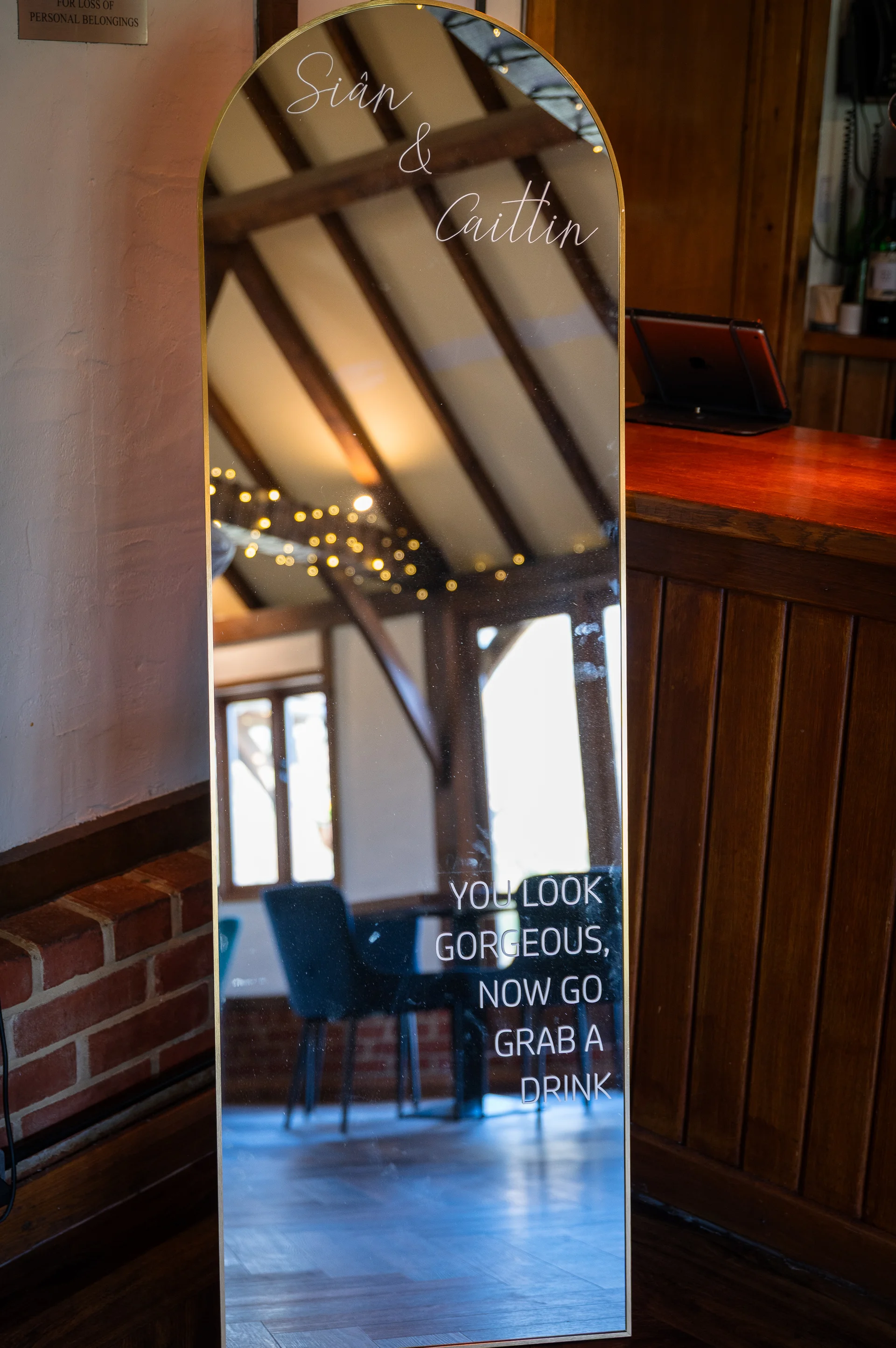 An arched full-length mirror featuring personalized white lettering that reads "Sian & Caitlin" and "You look gorgeous, now go grab a drink" reflects the rustic interior of a pub venue with exposed wooden beams and fairy lights.
