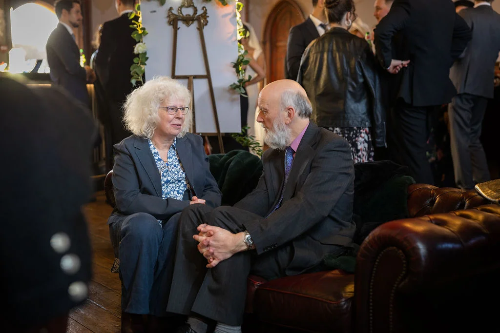 An older couple enjoys a quiet conversation while seated together on a leather sofa during the wedding reception.
