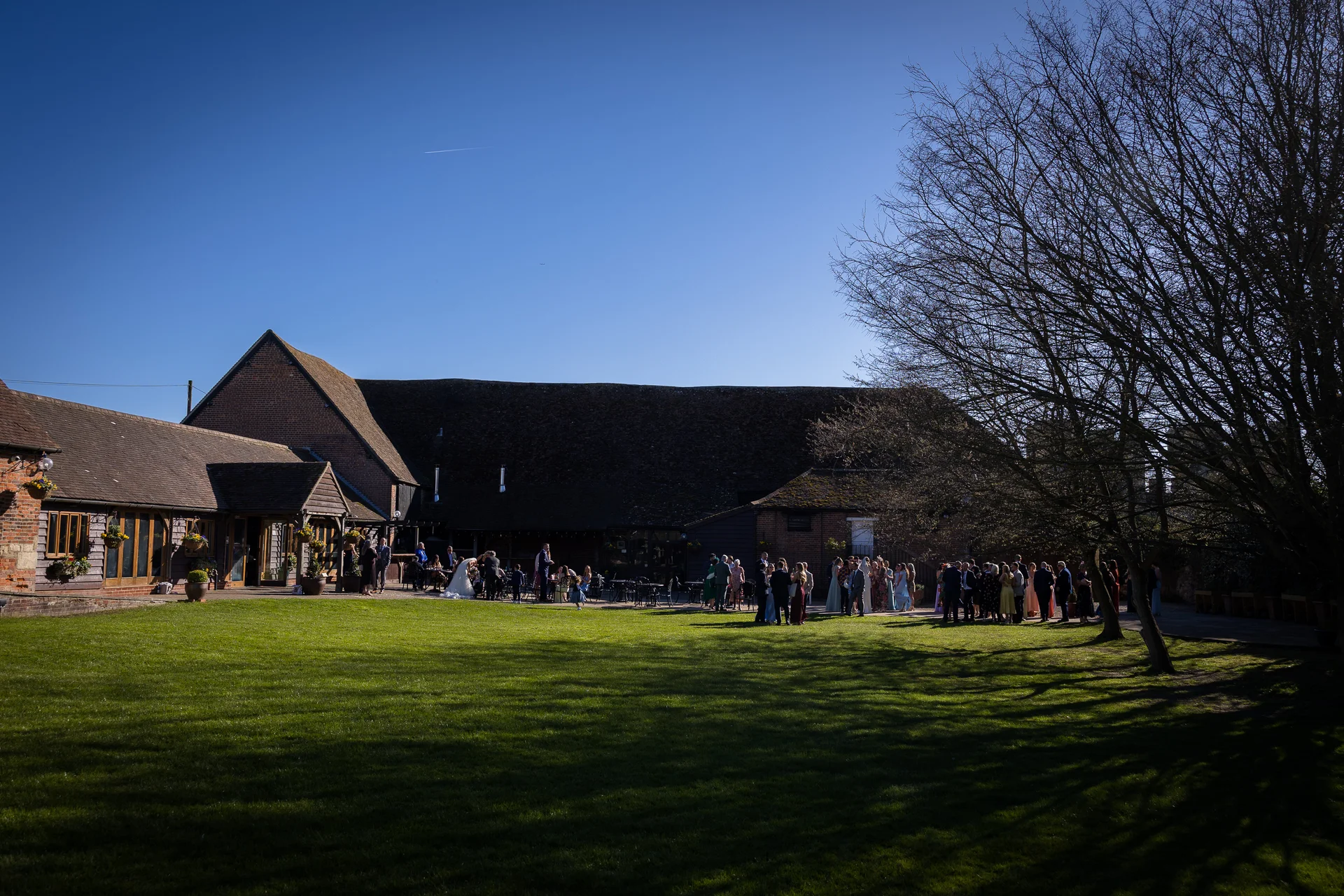 Guests mingle on a sunlit lawn outside a rustic barn wedding venue under a clear blue sky.