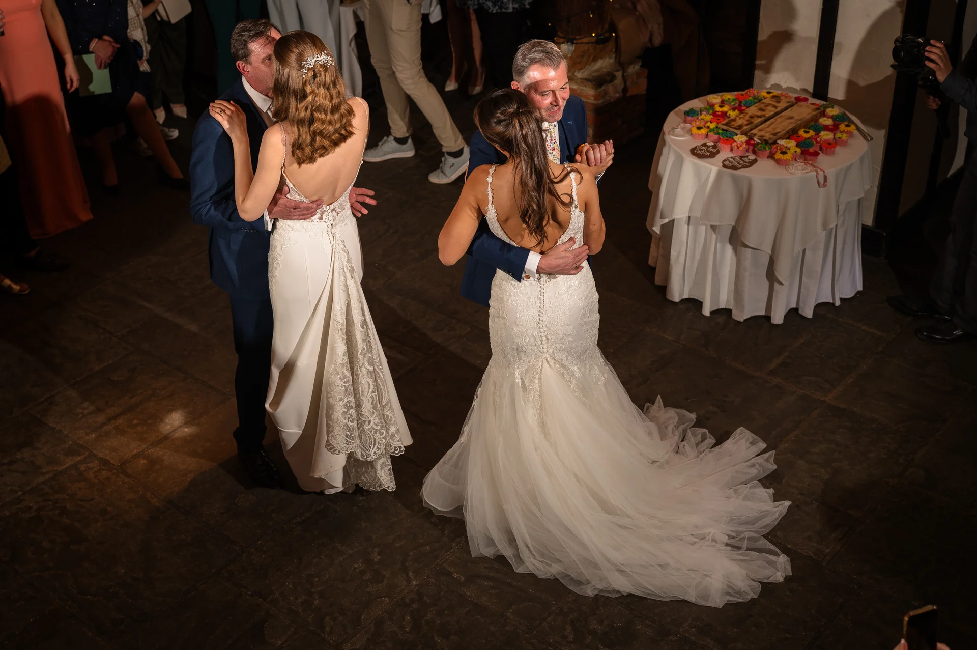 Two couples share a joyful dance on a stone floor during an evening wedding reception near a table displaying cupcakes.