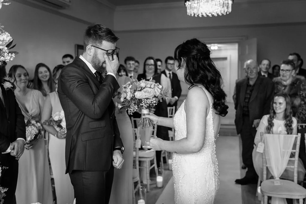 A tearful groom wipes away a tear at the altar during a black and white wedding ceremony while his bride holds her bouquet.