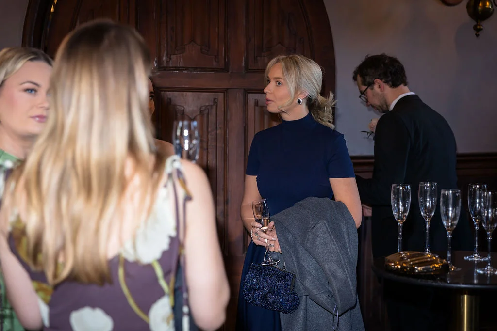 Guests mingling at a wedding drinks reception, with a blonde woman in a navy dress holding a glass of champagne and a dark beaded clutch bag.
