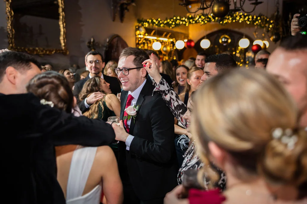 A groom wearing glasses and a tuxedo laughs joyfully among a crowded dance floor lit by warm fairy lights during a lively evening wedding reception.
