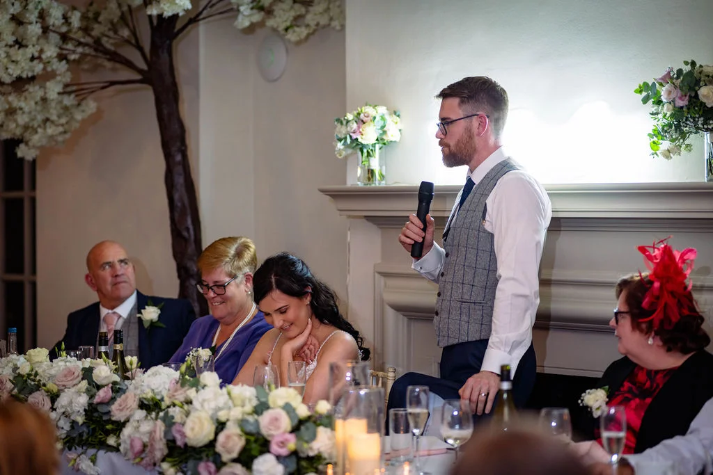 A groom delivers a speech while standing at the top table, with seated family members and the bride listening attentively amidst floral decorations.