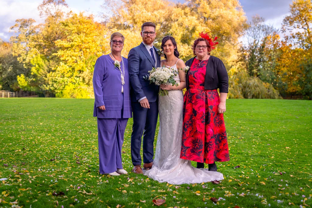 A newlywed couple poses outdoors on a lush green lawn with family members against a backdrop of vibrant autumn trees and golden foliage.