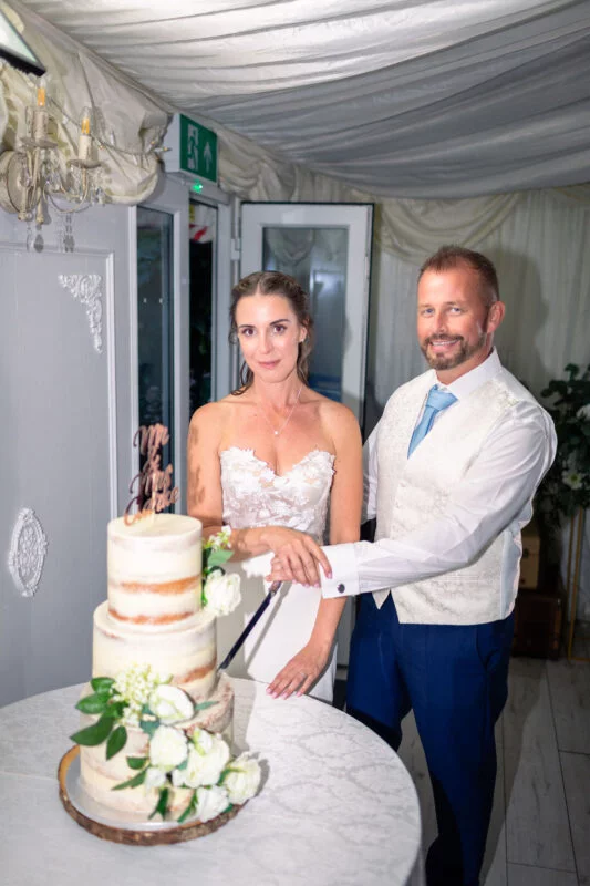 A smiling bride and groom stand together inside a draped marquee, holding a cake cutting knife over a three-tiered semi-naked wedding cake decorated with white flowers.